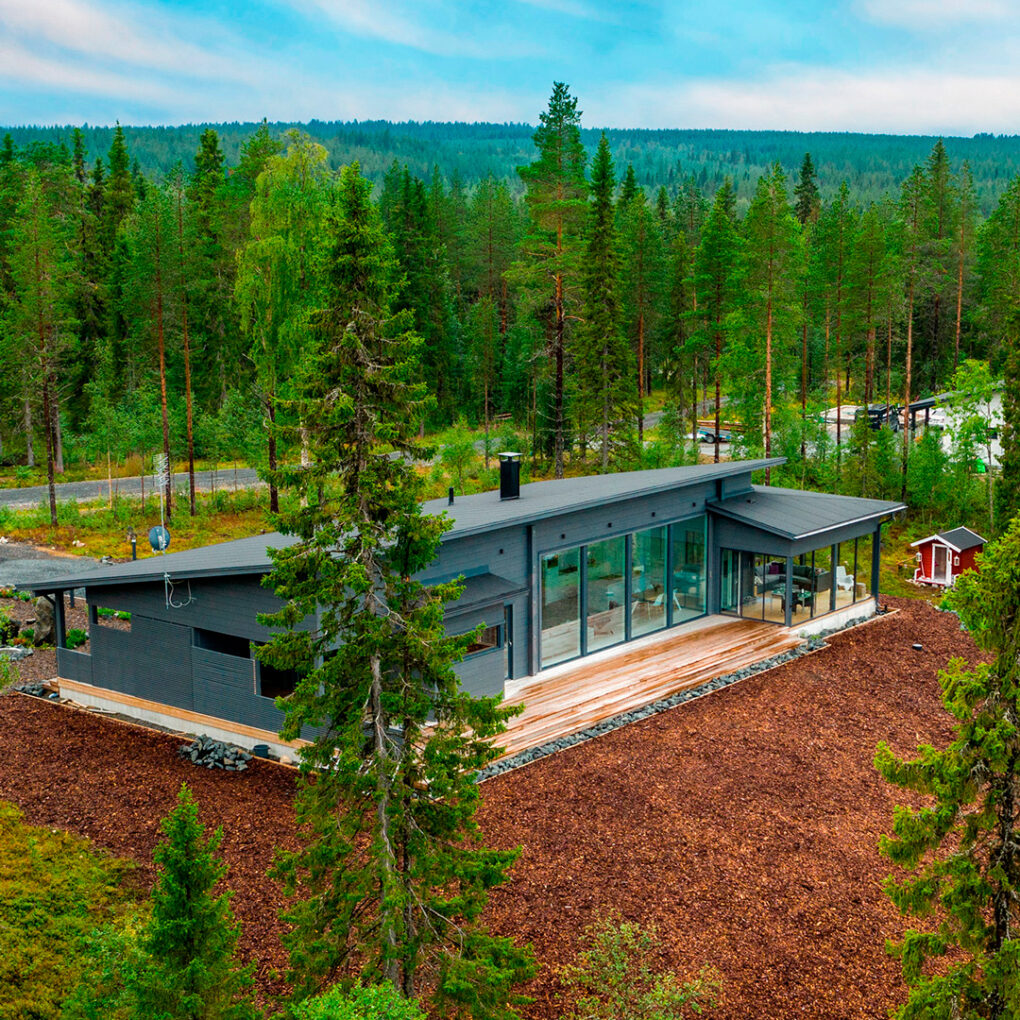 Pulpettikattoinen, harmaa hirsitalo metsän keskellä ilmasta kuvattuna.