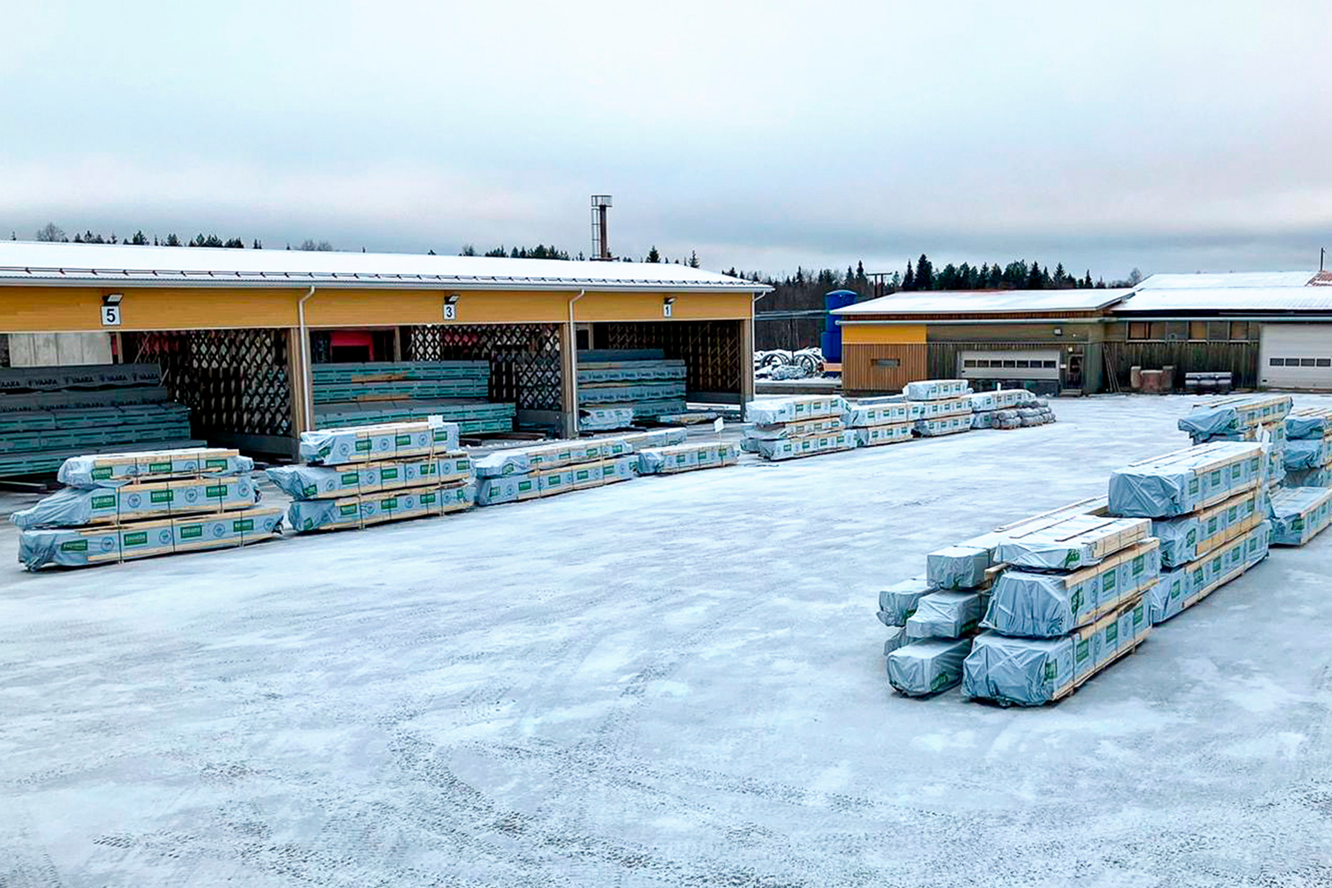 Talvinen kuva tehdasalueelta. Taka-alalla on varastokatos ja etualalla on kaksi riviä hirsi- ja puutavarapaketteja.