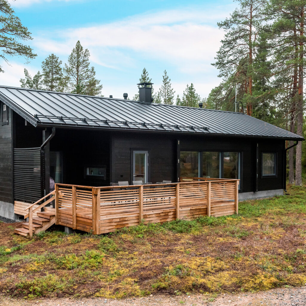 Musta harjakattoinen hirsihuvila, jonka etualalla on kivasti terassitilaa. Huvilaa ympäröi mäntymetsä.