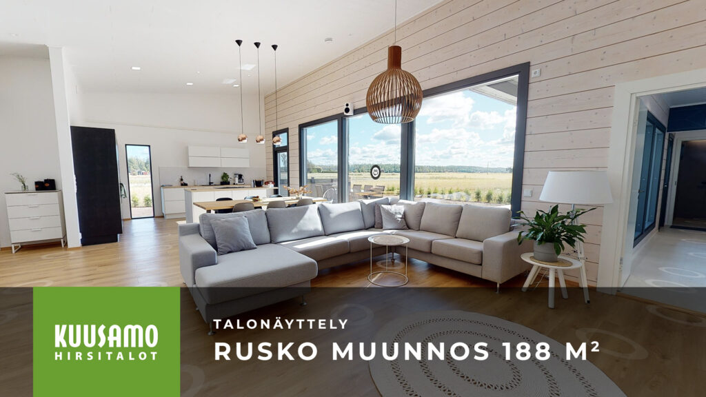 Esiteltävän hirsitalo Ruskon olohuoneessa on iso kulmasohva. Taustalla on vaaleilla väreillä sisustettu keittiö.