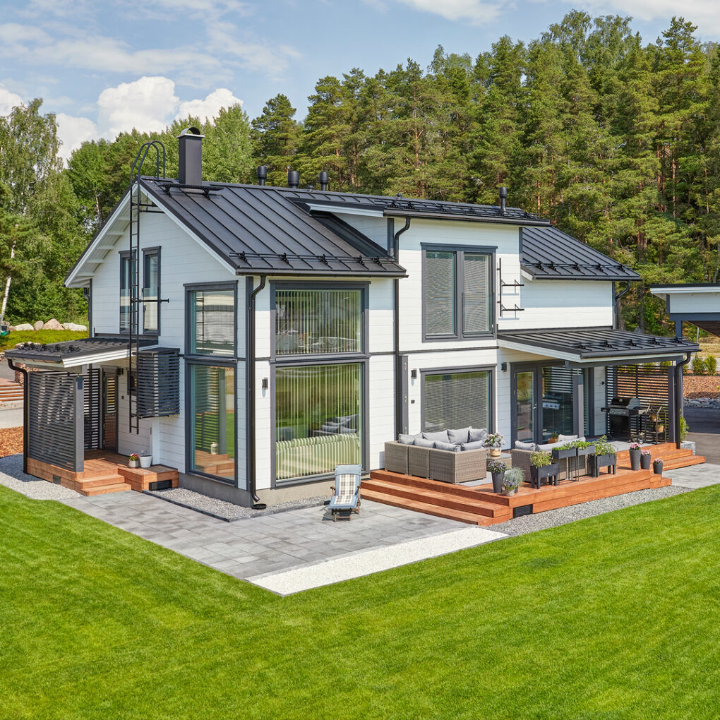 Kaksikerroksinen harjakattoinen hirsikoti. Valkoiseksi maalatussa Villa Hiisi -kodissa on harmaat vuorilaudat tuomassa kontrastia väritykseen.