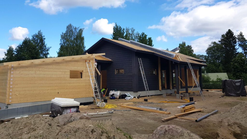 På bilden syns ett timmerhus och en carport. Timmerhuset är målat svart och carporten är ännu omålad. På gården finns byggavfall.