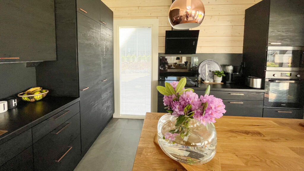 The kitchen units are black, and the handles are made of reddish metal. There is a beautiful bouquet of light pink flowers on the island.