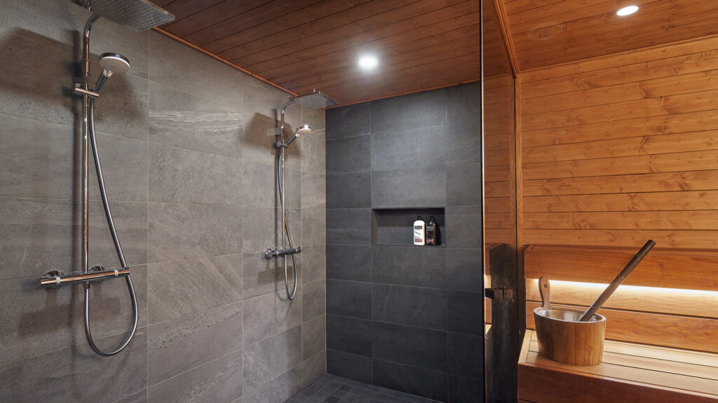 The washroom has dark grey tiles and the Sauna has dark brown wooden surfaces. The spaces are divided by a glass wall.