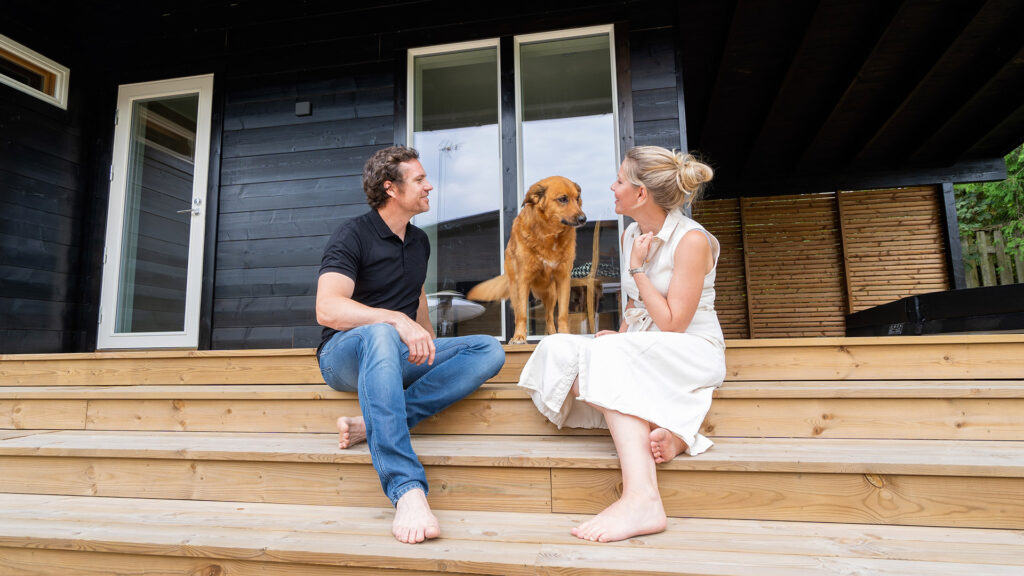 A couple sitting on the terrace of a log house with their dog.