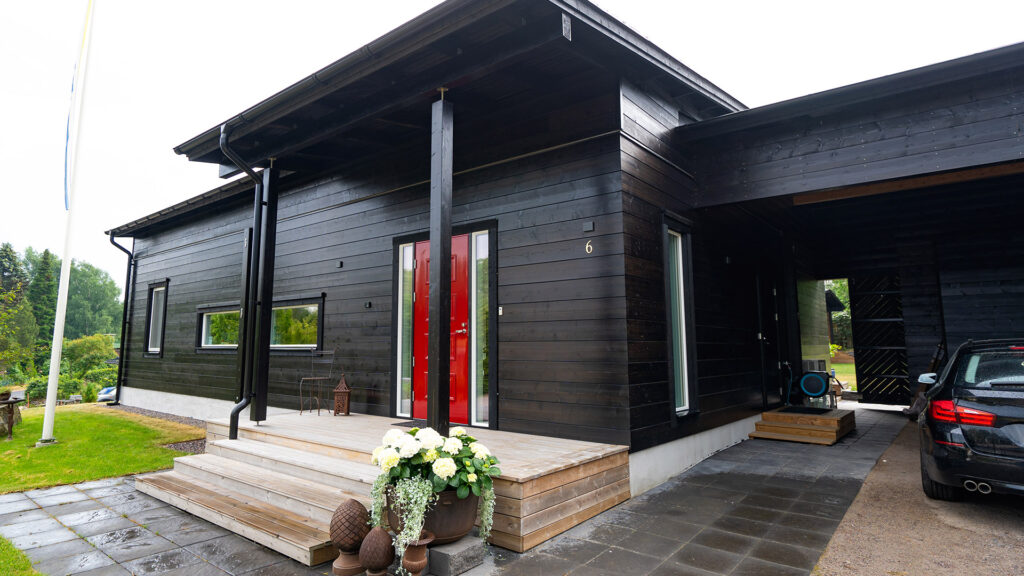 A black, modern log house with sheered corner joints. The red front door is a stylish detail on an otherwise black house.