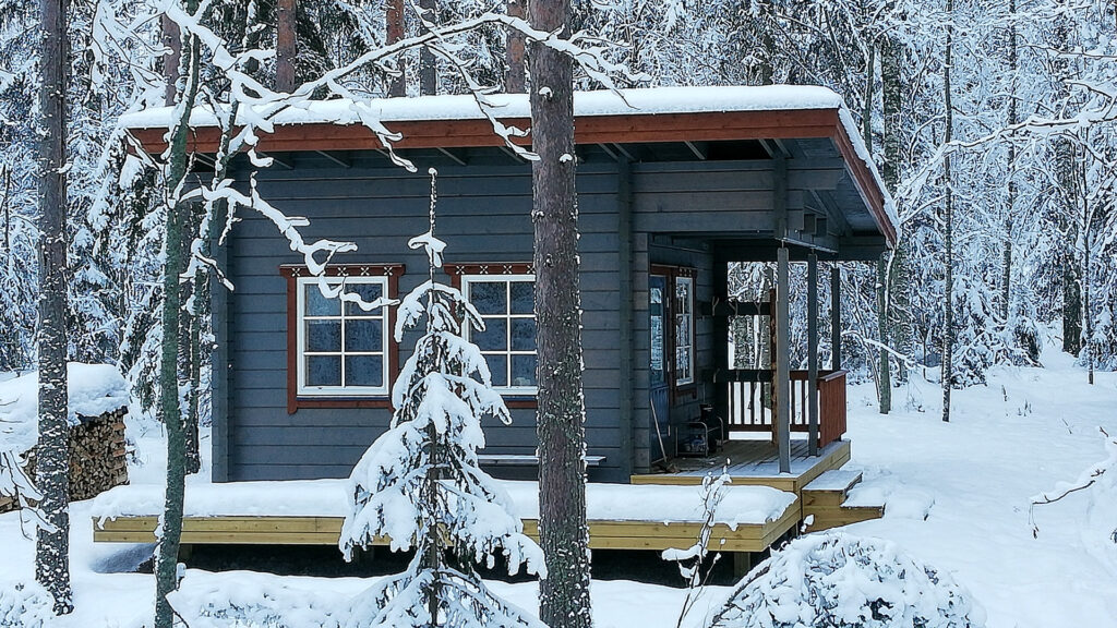 Pieni hirsiaitta lumisen metsämaiseman keskellä. Aitan edustalla on hieman terassitilaa.