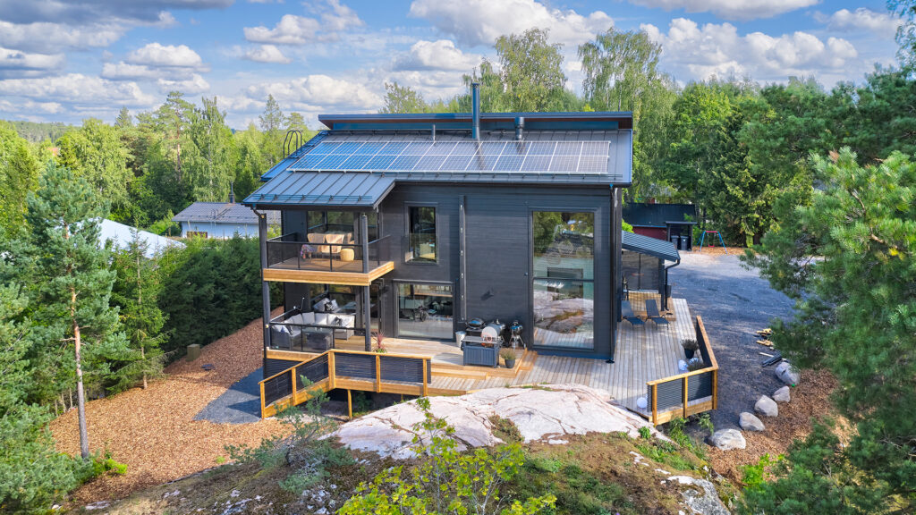 A two-storey modern log house featuring a hipped roof. The roof is fitted with solar panels and the house boasts plenty of terrace space.