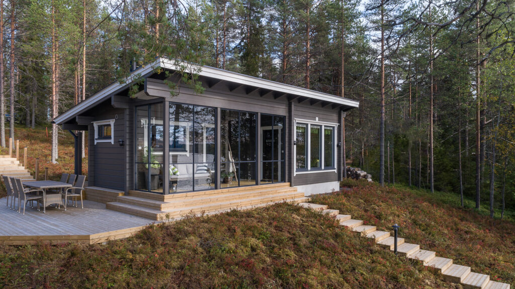 Saunamökki pulpettikatolla. Lasitettu terassi etualalla tuo lisätilaa oleskeluun. Rakennus on tehty rinteeseen.