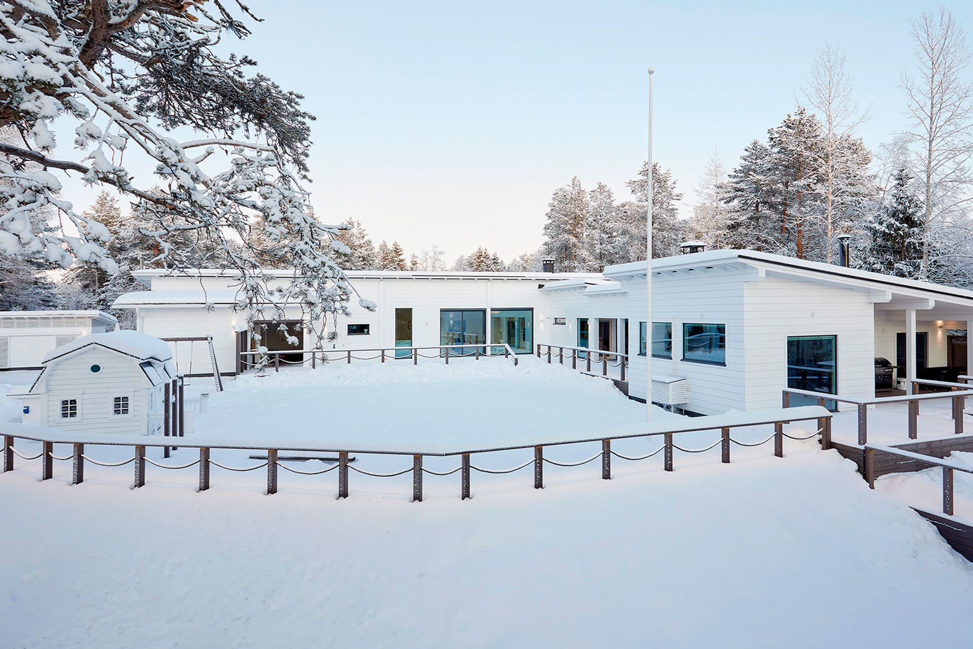 Valkoinen, L-mallinen ja pulpettikattoinen hirsitalo. Sen pihalla on myös pieni mansardikattoinen leikkimökki.