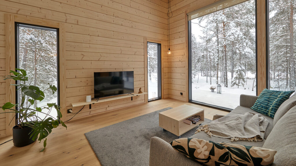 A living room with a sofa and a television. The living room features large windows that look out directly onto the woodland surrounding the house.