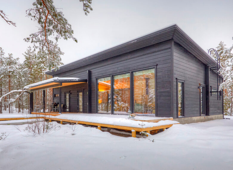 A black log house with a pitched roof. The orange light shining from inside the house lends a warm atmosphere to the otherwise wintry landscape.