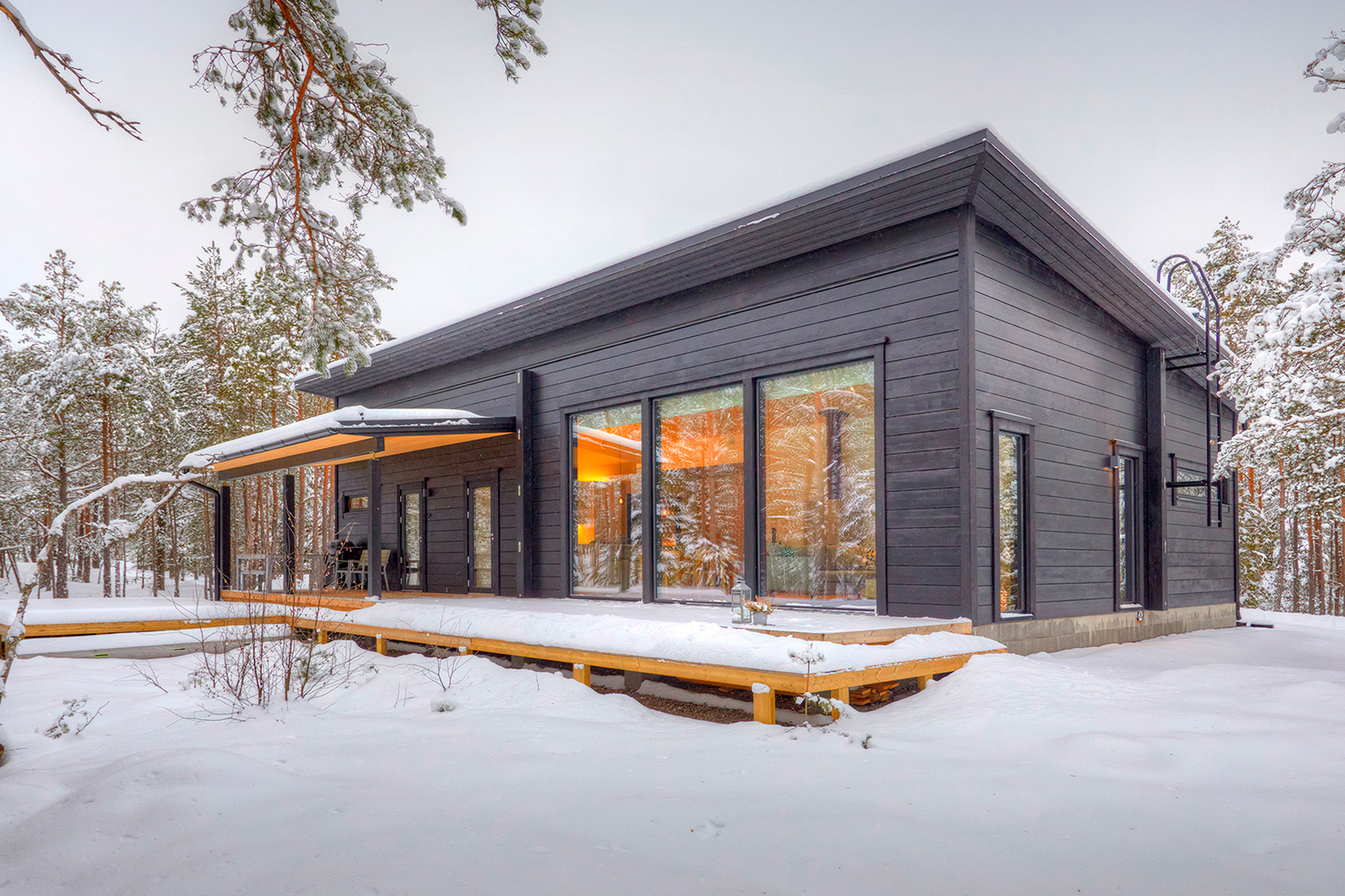 A black log house with a pitched roof. The orange light shining from inside the house lends a warm atmosphere to the otherwise wintry landscape.
