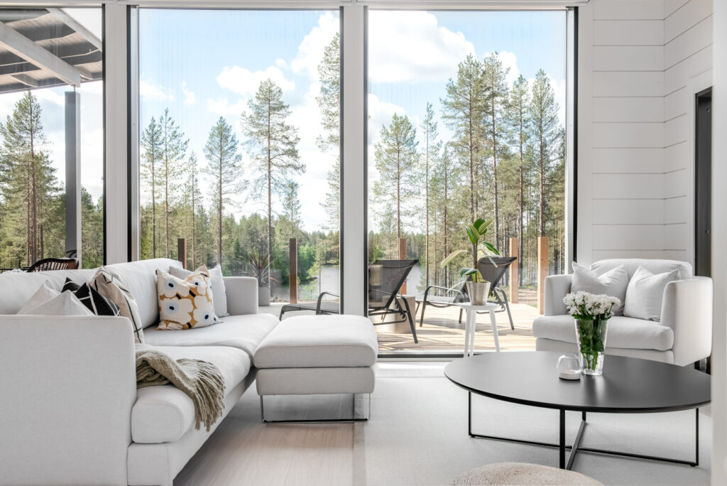 Valkoiseksi sisustettu hirsikodin olohuone. Suurista ikkunoista avautuu maisemat läheiseen vesistöön. Talon vierestä alkaa heti mäntymetsä.