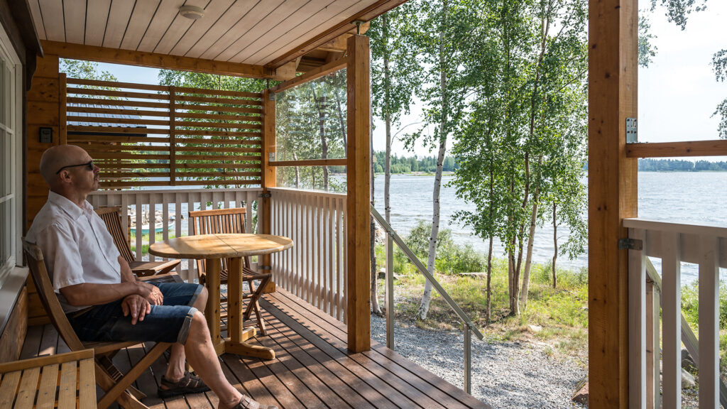 Mökin omistaja nauttii kesäpäivästä mökkinsä terassilla istuskellen. Edessä aukeaa merimaisema.