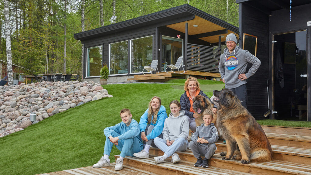 Heleniuksen perhe modernin hirsimökkinsä edustalla. Perheeseen kuuluu vanhemmat, neljä lasta sekä kaksi koiraa.