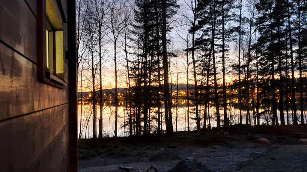 Iltahämärässä otettu kuva. Vasemmalla näkyy hirsitalon seinää ja metsän takaa pilkistää kaupungin valot.