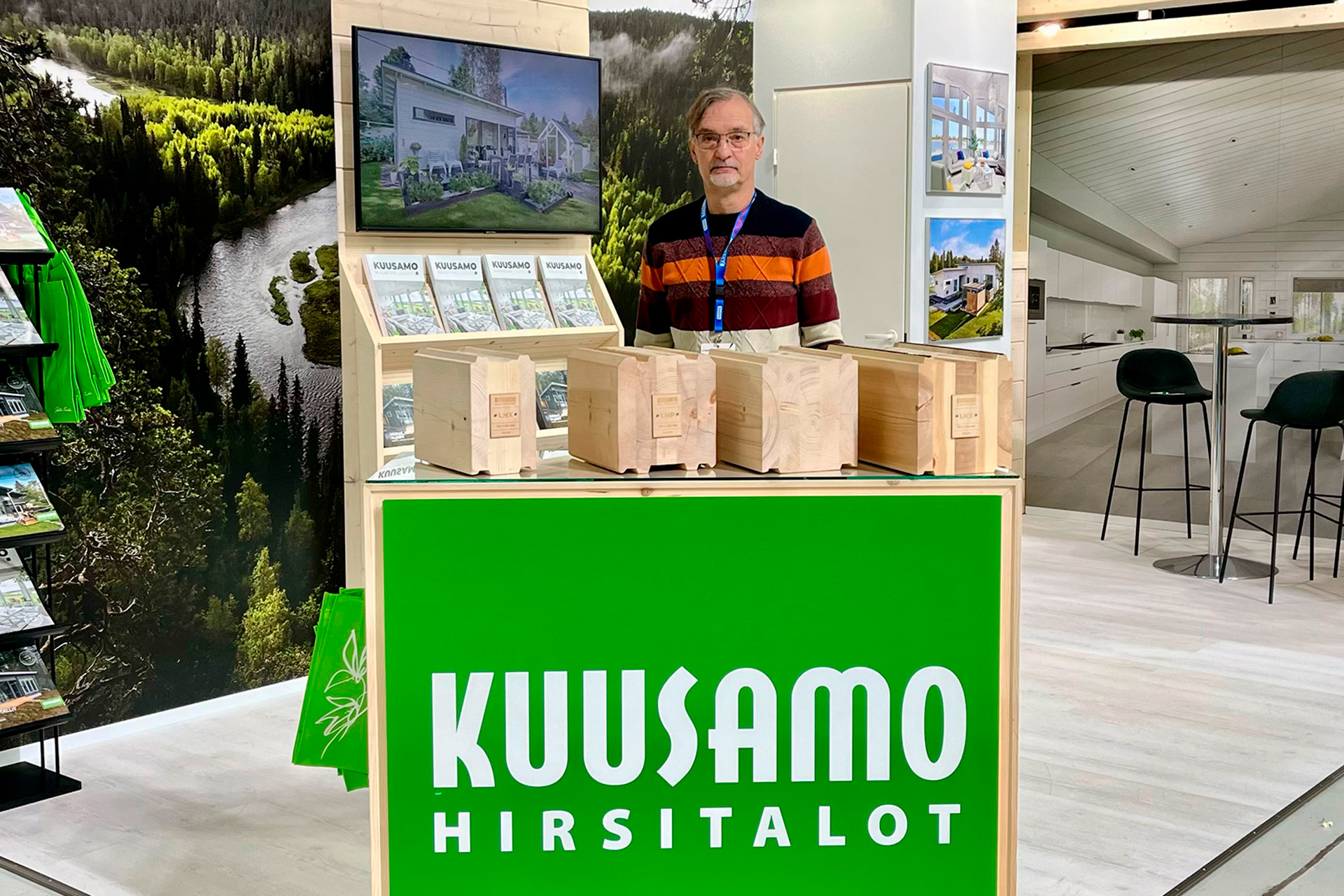 Kuusamo-myyjä Ari Hottinen messuosastolla Kuopiossa. Osastolla on tarjolla esitteitä ja näytillä on sen lisäksi hirsien mallipaloja.