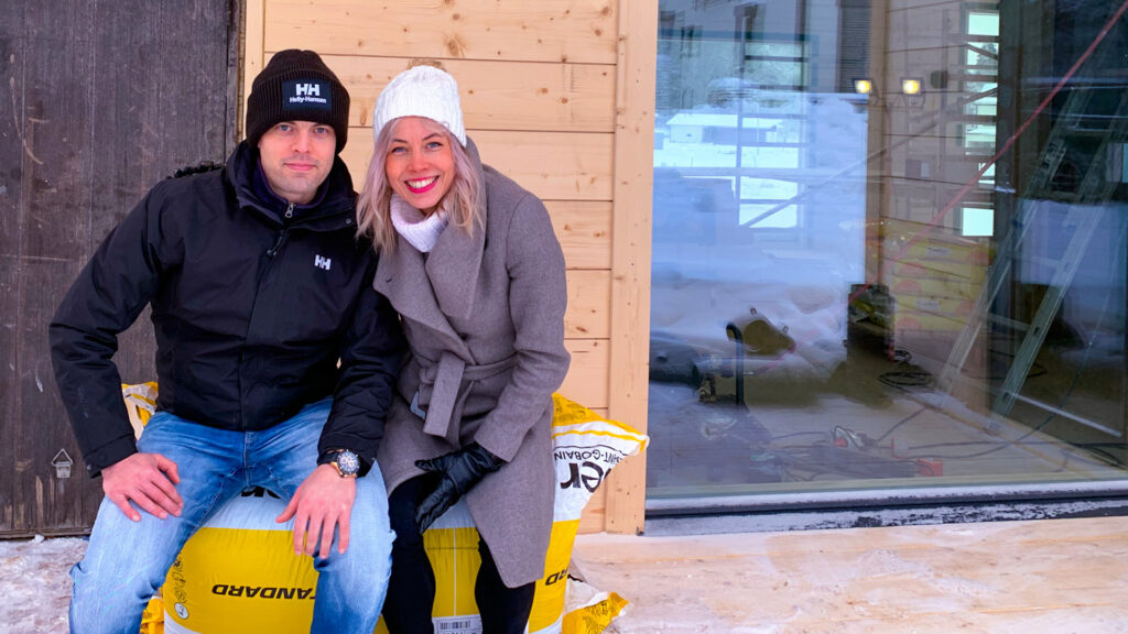 Johanna och Henkka sitter på ett paket med isolering framför sitt timmerhus som är under uppförande.