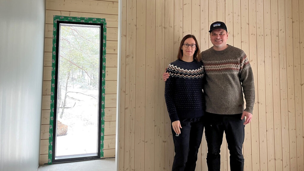 A couple in a log house under construction, photographed in front of a vertical-panelled partition wall.