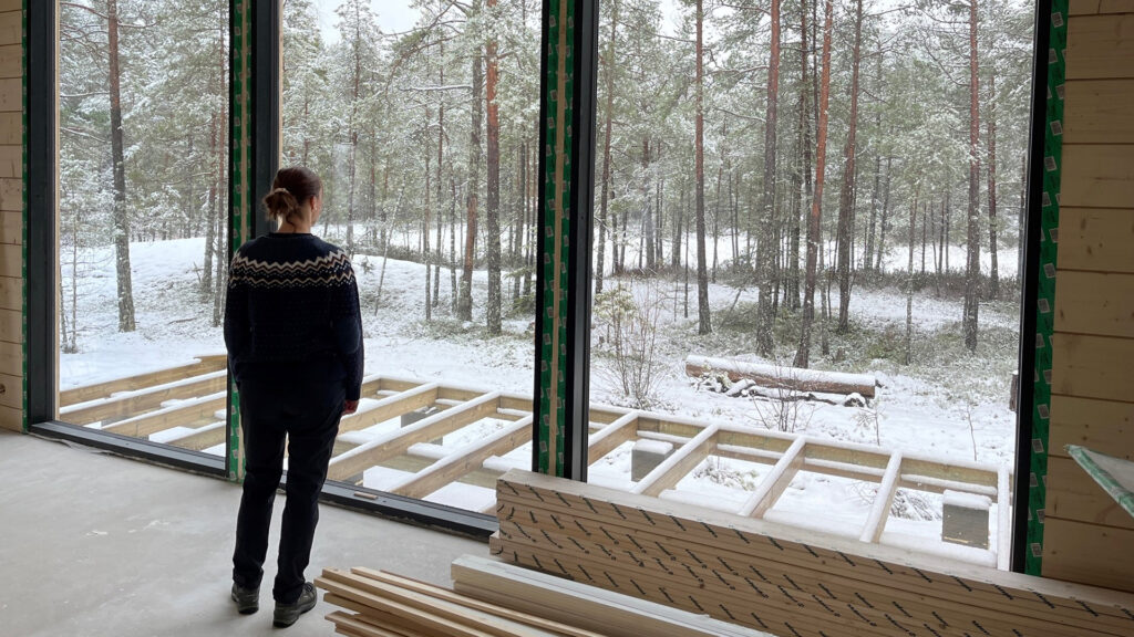 A woman looks out of the large windows of the log house under construction towards the nearby forest.