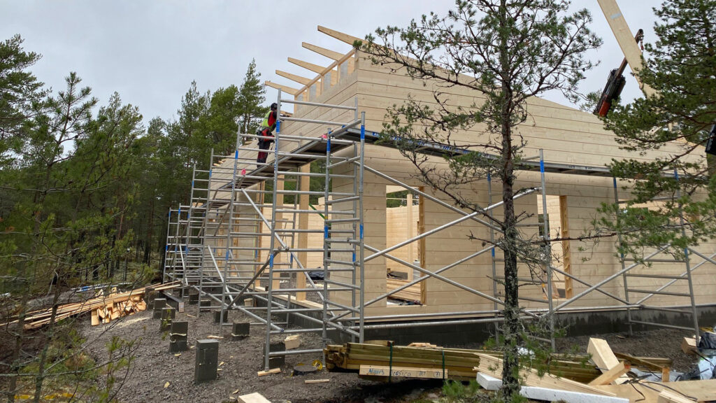 A photo of the log house being erected. A crane is lifting a ridge beam onto the roof. Five ridge beams are already in place.