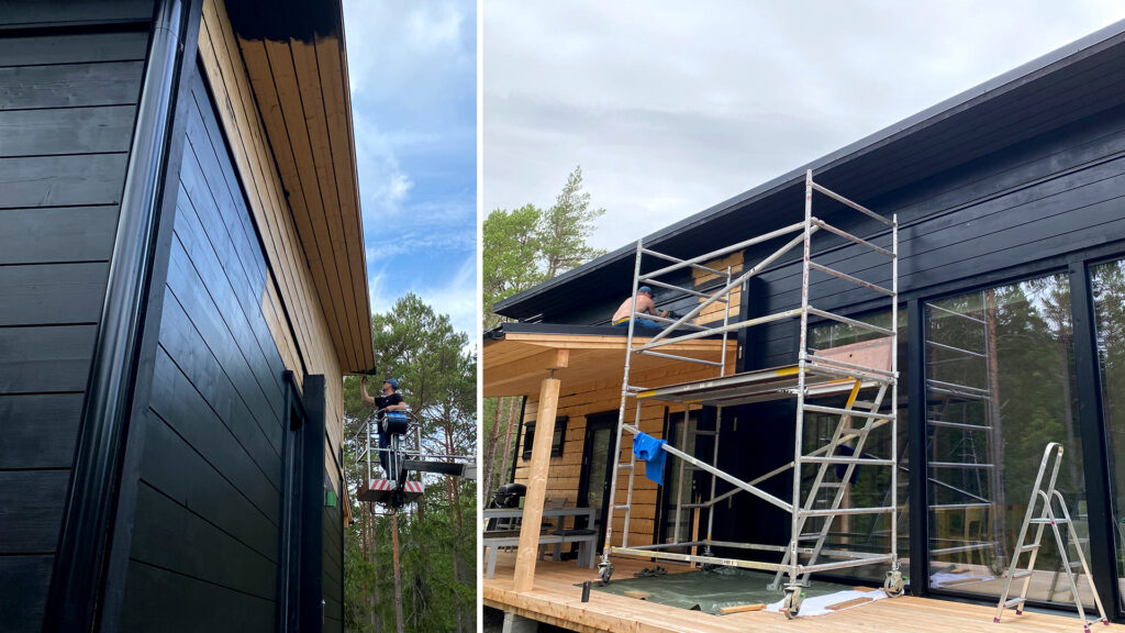 A log house is being painted black. Scaffolding and a man-lift are being used to assist with the work.