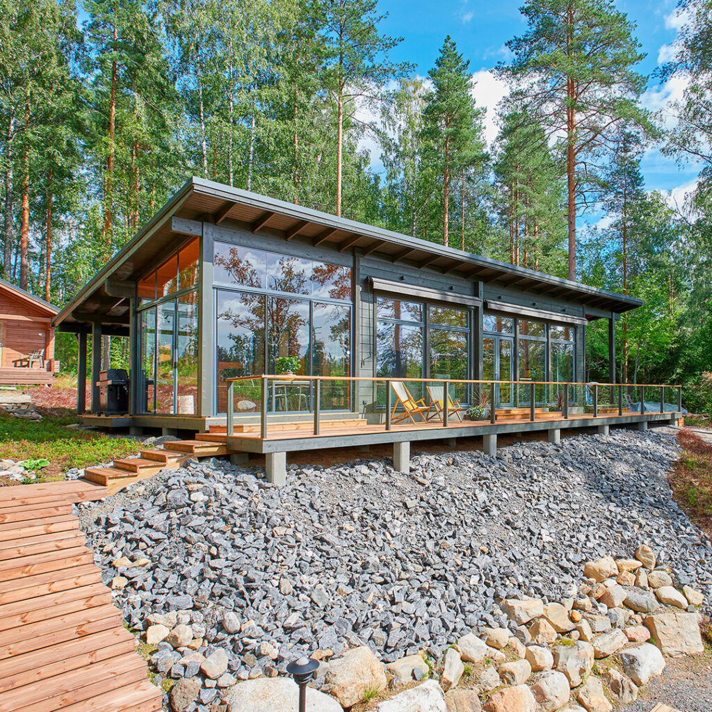 Pulpettikattoinen moderni hirsihuvila, jonka päädyssä on lasitettu terassi. Huvilan edessä olevalta terassilta lähtee portaat kohti rantaa.