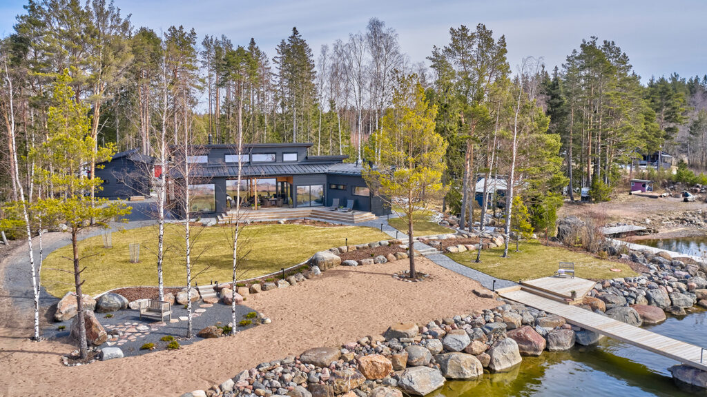 Hirsitalo ja sen pihapiiri kuvattuna yläilmoista. Pihasuunnittelussa on tasoja. Rannassa on isoja kiviä, sitten tulee hiekkaa ja juuri ennen taloa on nurmialue.