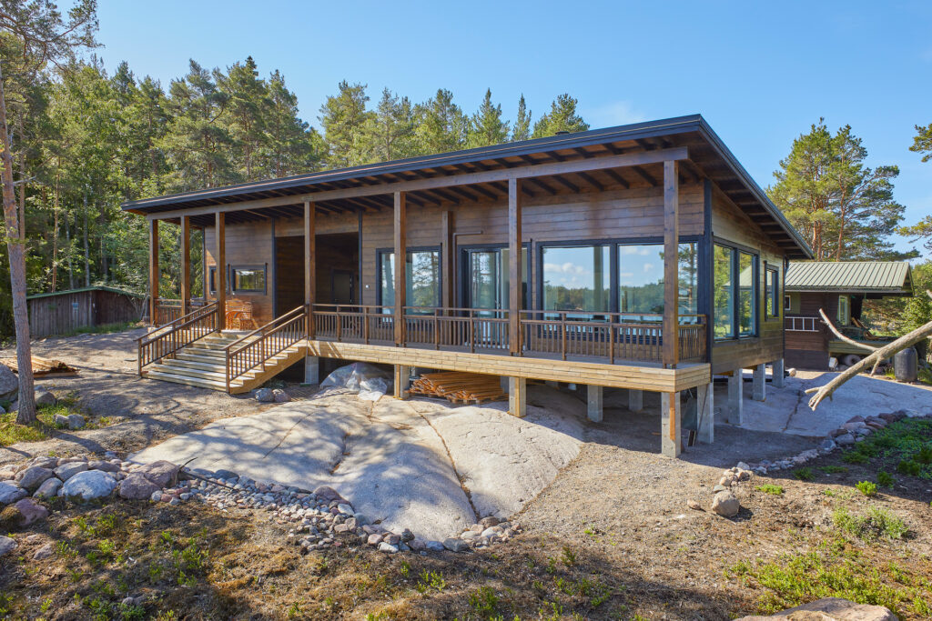 Pulpettikattoinen pitkän mallinen hirsihuvila kalliolla. Takana on mäntymetsää.