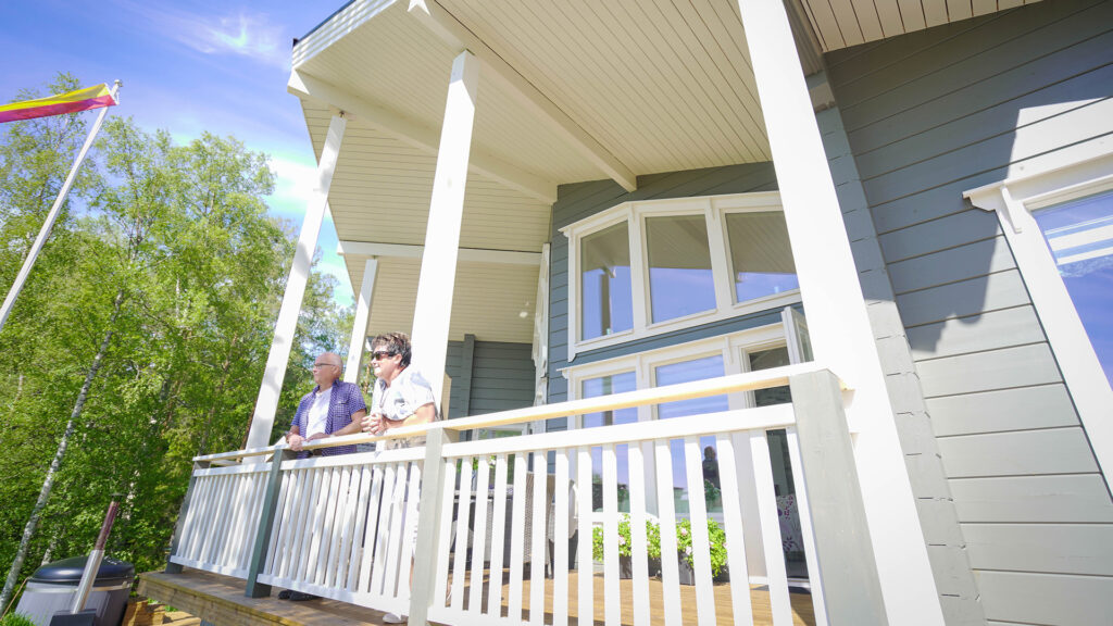 A couple on the terrace of their Nuuna log house in sunny weather. A flag is flying from the flagpole.