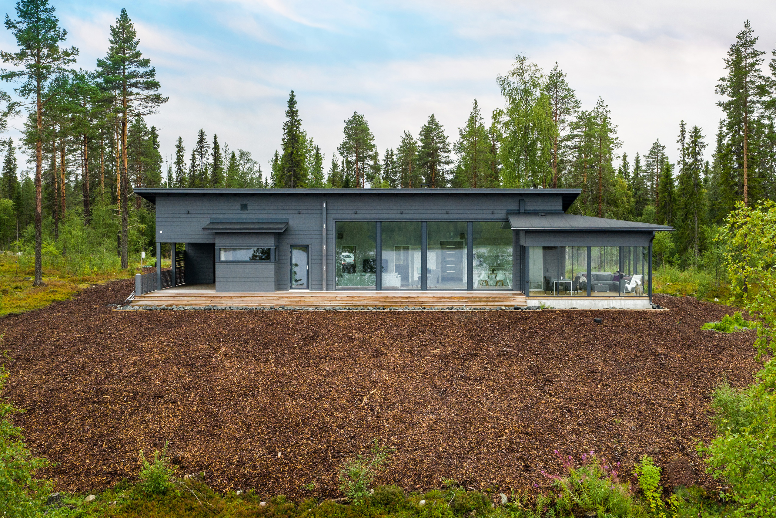 Tummanharmaa, pulpettikattoinen hirsitalo. Hirsitalon yhteydessä on lasitettu terassi.