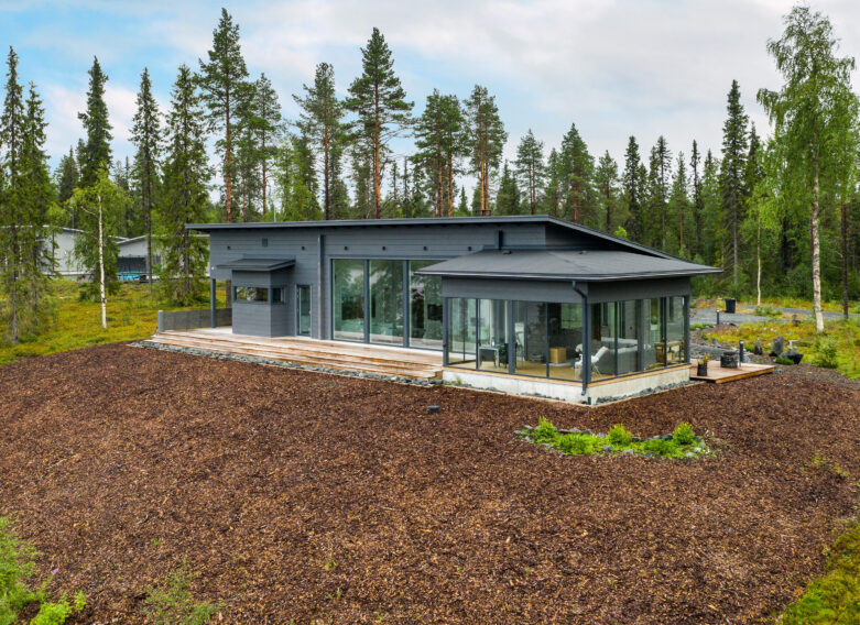 Tummanharmaa, pulpettikattoinen hirsitalo suomalaisen metsämaiseman keskellä.