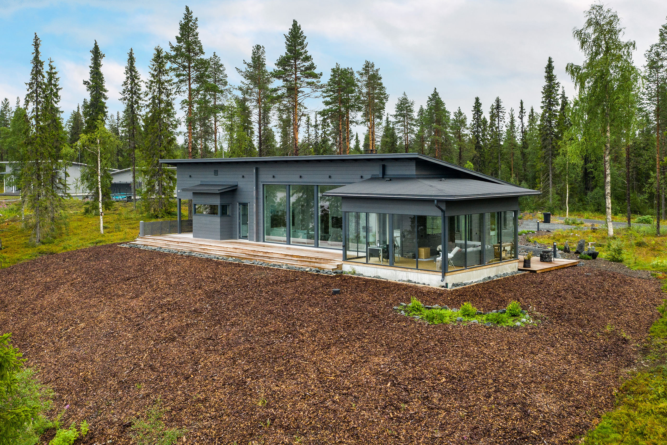 Tummanharmaa, pulpettikattoinen hirsitalo suomalaisen metsämaiseman keskellä.